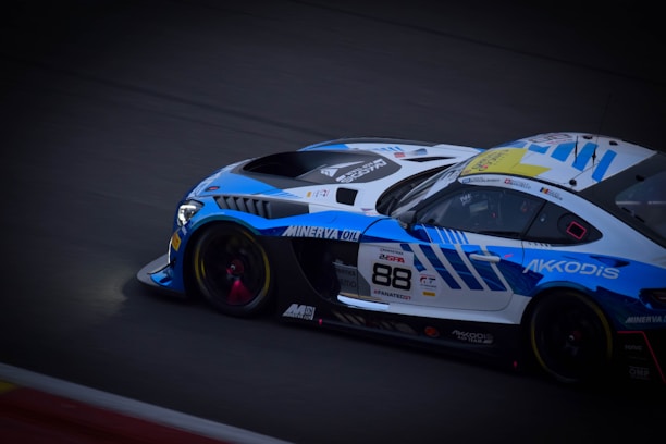 A sleek, high-performance racing car with a vibrant blue and white color scheme is captured in motion on a track. The car features branding logos such as Minerva Oil and Akkodis, and the number 88 is prominently displayed on the side. Design elements suggest it is equipped for professional racing, with aerodynamic features and sponsorship decals visible.