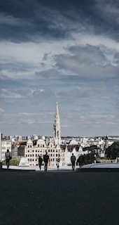 A gothic-style building with a tall spire stands prominently in the middle of an urban landscape. The foreground features several people walking on a dark path leading towards the cityscape. The sky is partly cloudy, casting a moody and overcast vibe.