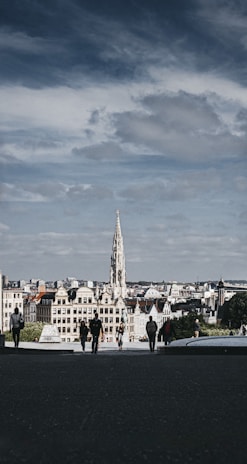 A gothic-style building with a tall spire stands prominently in the middle of an urban landscape. The foreground features several people walking on a dark path leading towards the cityscape. The sky is partly cloudy, casting a moody and overcast vibe.