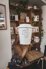 A hand holding a white to-go coffee cup with the label 'My Little Cup' in a cozy cafe setting. The background features wooden shelves adorned with plants and various items, contributing to a warm, inviting atmosphere.