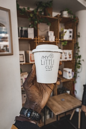 A hand holding a white to-go coffee cup with the label 'My Little Cup' in a cozy cafe setting. The background features wooden shelves adorned with plants and various items, contributing to a warm, inviting atmosphere.