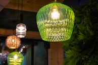 Several decorative hanging lamps are illuminated, with a prominent green glass lamp in the foreground featuring a textured pattern. Other lamps are visible in the background, including a woven one and a clear glass one. The setting includes lush greenery on the right side and wooden elements, creating a cozy atmosphere.