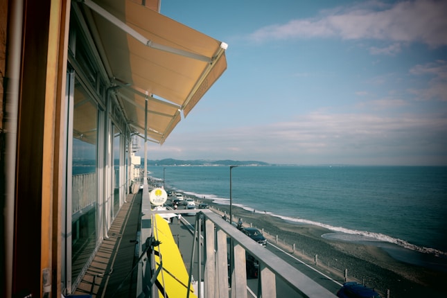 A coastal scene with a view of the ocean and a wooden boardwalk alongside a building with glass windows and yellow awnings. The sky is partly cloudy, and there is a railing along the walkway. Cars are parked along a road that runs parallel to the shoreline.