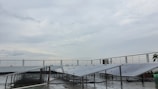 A rooftop setup with several solar panels positioned at an angle. The panels are supported on metal frames above a wet surface, suggesting recent rain. A railing surrounds the rooftop area, and the sky is overcast with gray clouds.