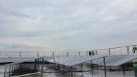 A rooftop setup with several solar panels positioned at an angle. The panels are supported on metal frames above a wet surface, suggesting recent rain. A railing surrounds the rooftop area, and the sky is overcast with gray clouds.