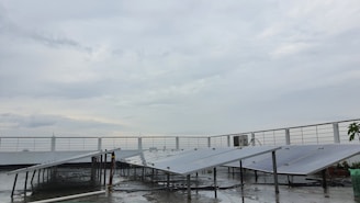 A rooftop setup with several solar panels positioned at an angle. The panels are supported on metal frames above a wet surface, suggesting recent rain. A railing surrounds the rooftop area, and the sky is overcast with gray clouds.