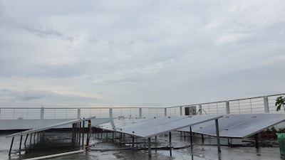 A rooftop setup with several solar panels positioned at an angle. The panels are supported on metal frames above a wet surface, suggesting recent rain. A railing surrounds the rooftop area, and the sky is overcast with gray clouds.
