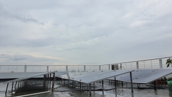 A rooftop setup with several solar panels positioned at an angle. The panels are supported on metal frames above a wet surface, suggesting recent rain. A railing surrounds the rooftop area, and the sky is overcast with gray clouds.
