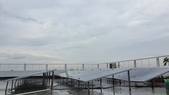 A rooftop setup with several solar panels positioned at an angle. The panels are supported on metal frames above a wet surface, suggesting recent rain. A railing surrounds the rooftop area, and the sky is overcast with gray clouds.