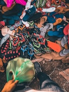 A variety of flip-flops and sandals in multiple colors and styles are scattered on a surface. The shot includes a person wearing jeans and a black shoe standing near the footwear, holding a green plastic bag filled with items.