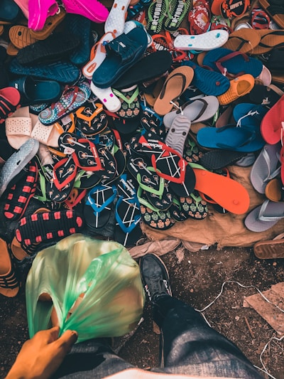 A variety of flip-flops and sandals in multiple colors and styles are scattered on a surface. The shot includes a person wearing jeans and a black shoe standing near the footwear, holding a green plastic bag filled with items.