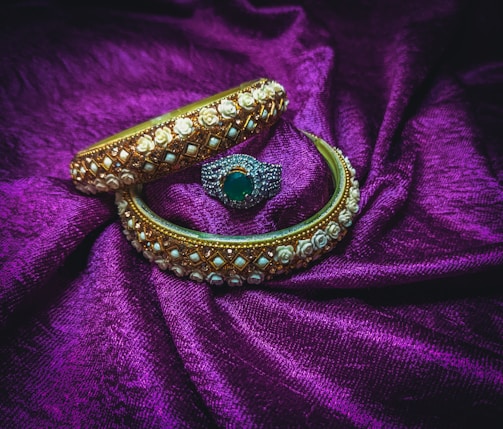 Two ornate bangles and a ring are placed on a rich purple fabric. The bangles are decorated with intricate gold and white floral designs, while the ring features a central green gemstone surrounded by small sparkling stones.