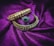 Two ornate bangles and a ring are placed on a rich purple fabric. The bangles are decorated with intricate gold and white floral designs, while the ring features a central green gemstone surrounded by small sparkling stones.