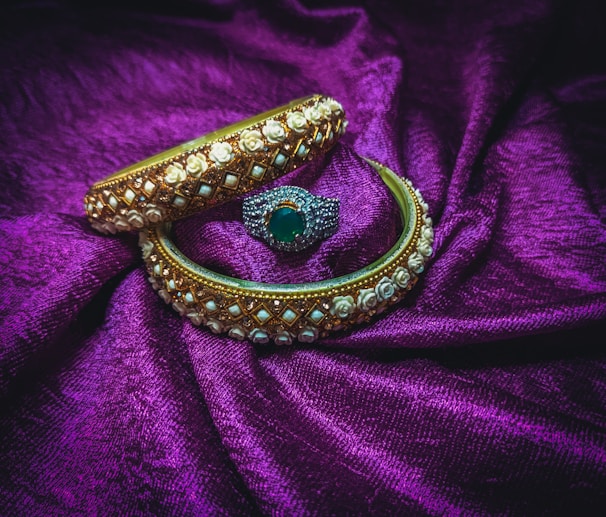 Two ornate bangles and a ring are placed on a rich purple fabric. The bangles are decorated with intricate gold and white floral designs, while the ring features a central green gemstone surrounded by small sparkling stones.