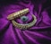 Two ornate bangles and a ring are placed on a rich purple fabric. The bangles are decorated with intricate gold and white floral designs, while the ring features a central green gemstone surrounded by small sparkling stones.