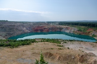 A large open-pit mine with steep, terraced walls surrounds a body of turquoise water. The pit is situated in a landscape with sparse vegetation and patches of greenery, with a dense forest visible in the background under a clear blue sky.