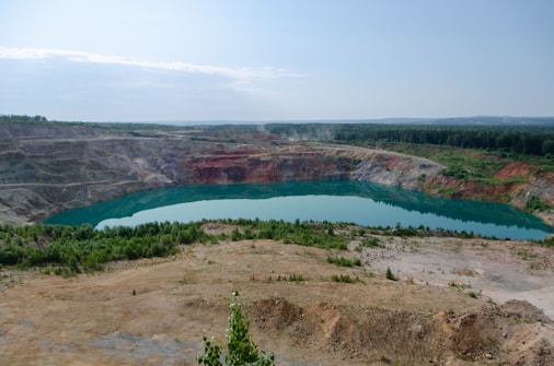 A large open-pit mine with steep, terraced walls surrounds a body of turquoise water. The pit is situated in a landscape with sparse vegetation and patches of greenery, with a dense forest visible in the background under a clear blue sky.