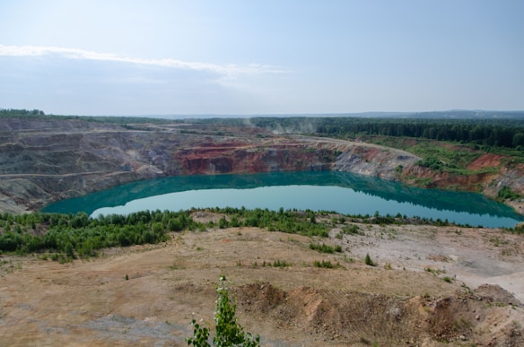 A large open-pit mine with steep, terraced walls surrounds a body of turquoise water. The pit is situated in a landscape with sparse vegetation and patches of greenery, with a dense forest visible in the background under a clear blue sky.