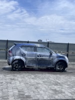 A car covered in soap and water appears to be undergoing a wash in an open area under a partly cloudy sky. The background includes a metal fence and some distant buildings, including a crane.