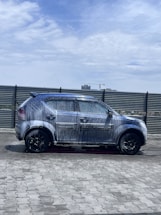 A car covered in soap and water appears to be undergoing a wash in an open area under a partly cloudy sky. The background includes a metal fence and some distant buildings, including a crane.