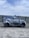 A car covered in soap and water appears to be undergoing a wash in an open area under a partly cloudy sky. The background includes a metal fence and some distant buildings, including a crane.