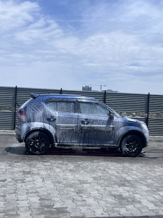 A car covered in soap and water appears to be undergoing a wash in an open area under a partly cloudy sky. The background includes a metal fence and some distant buildings, including a crane.