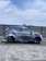A car covered in soap and water appears to be undergoing a wash in an open area under a partly cloudy sky. The background includes a metal fence and some distant buildings, including a crane.