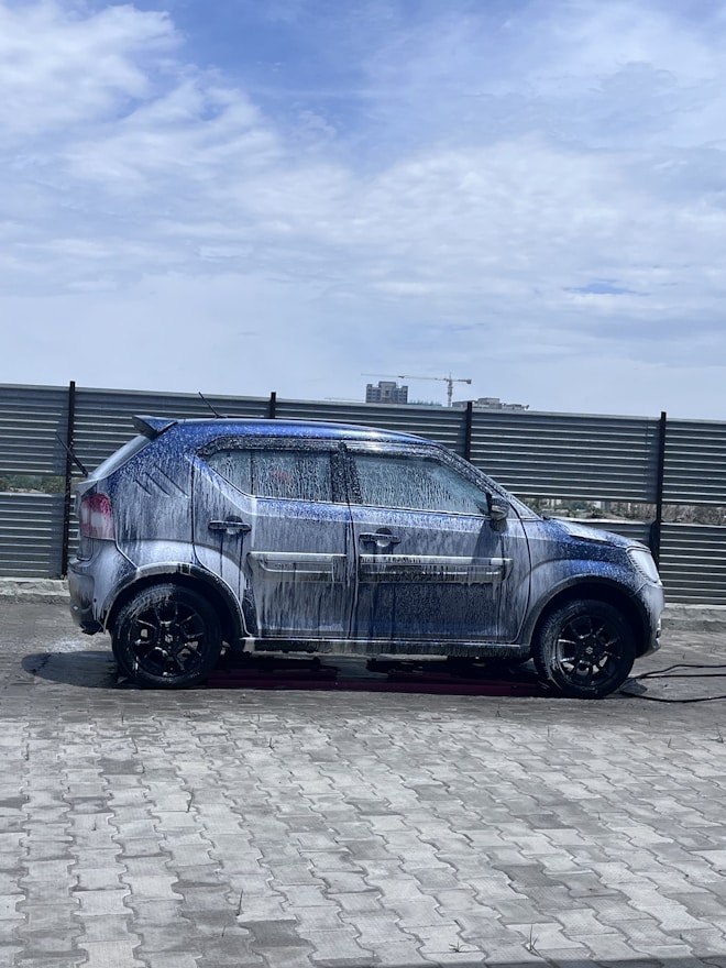 A car covered in soap and water appears to be undergoing a wash in an open area under a partly cloudy sky. The background includes a metal fence and some distant buildings, including a crane.
