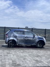 A car covered in soap and water appears to be undergoing a wash in an open area under a partly cloudy sky. The background includes a metal fence and some distant buildings, including a crane.