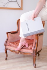 A classic upholstered chair with wooden frame features intricate detailing and a plush cushion in a soft blush color. A person is seated on the arm of the chair, wearing light-colored pants and pink sandals. The individual is holding a book titled 'I AM A CHAIR'. The setting is indoors with a wooden floor and a partially visible framed artwork on the wall.