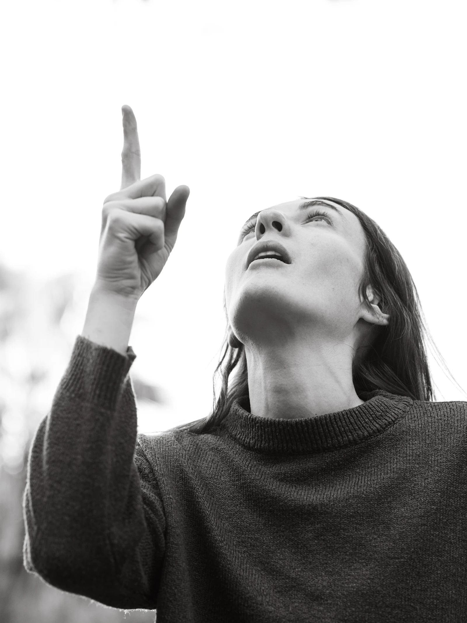A woman in black and white photography looks up toward the sky with her hand raised, a gesture of reaching for someone no longer there.
