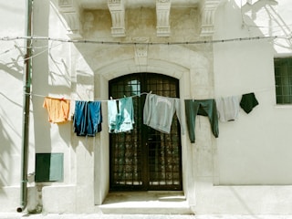 Clothes hang from a line strung across the facade of an old stone building. The building features a large arched doorway with metal bars and decorative stonework above. The hanging clothes, including shirts and shorts in various colors, sway gently in the breeze, casting soft shadows on the wall.