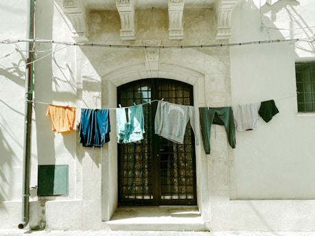 Clothes hang from a line strung across the facade of an old stone building. The building features a large arched doorway with metal bars and decorative stonework above. The hanging clothes, including shirts and shorts in various colors, sway gently in the breeze, casting soft shadows on the wall.
