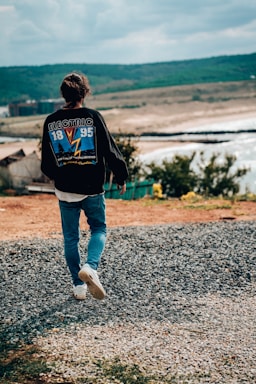 A person wearing a black jacket with a graphic design on the back, featuring the word 'Electric' and numbers '18' and '95', walking on a gravel path towards a scenic landscape. The background includes rolling hills, a shoreline, and a body of water under a cloudy sky.
