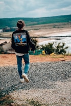 A person wearing a black jacket with a graphic design on the back, featuring the word 'Electric' and numbers '18' and '95', walking on a gravel path towards a scenic landscape. The background includes rolling hills, a shoreline, and a body of water under a cloudy sky.