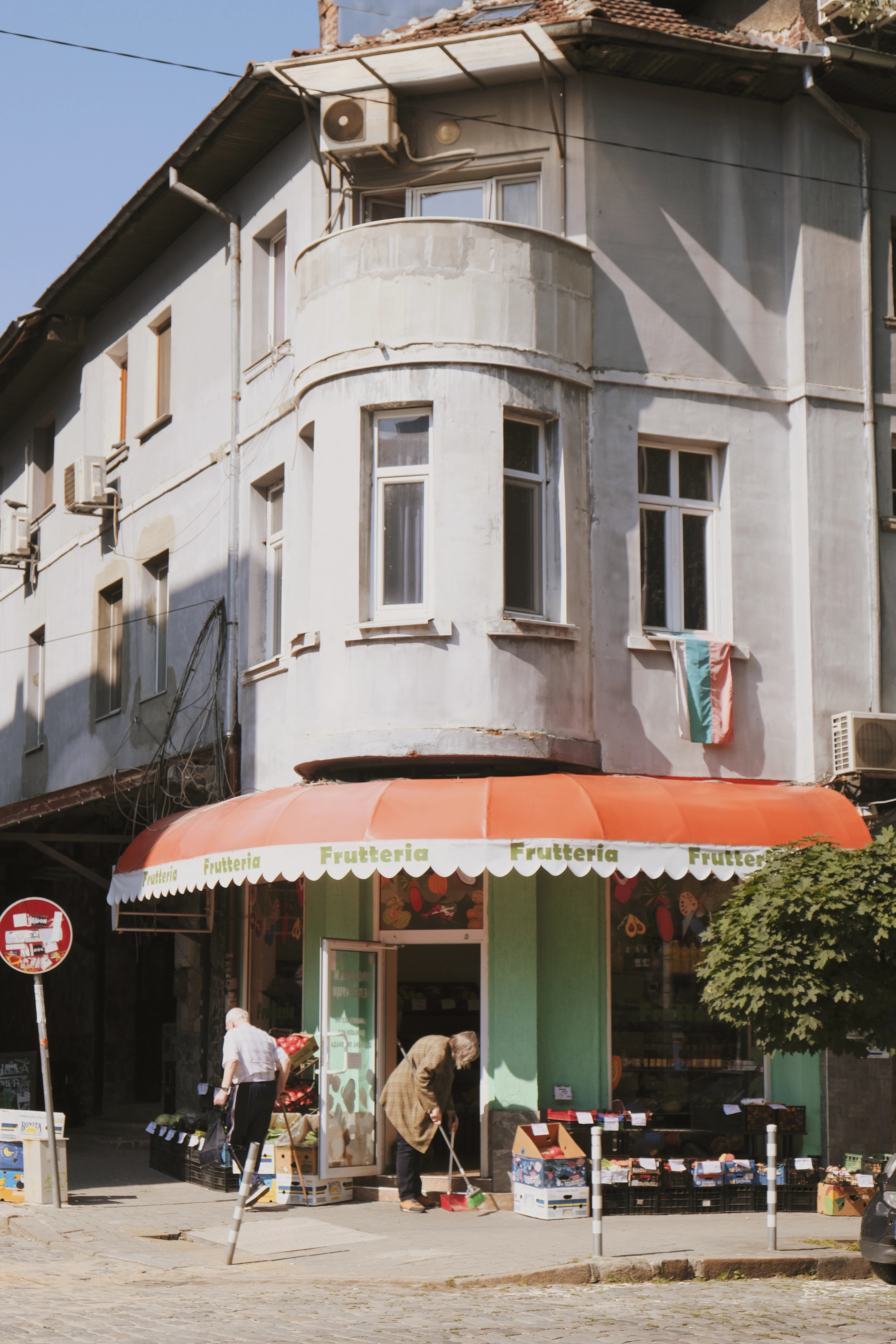 A corner greengrocer in Sofia beneath a striped orange awning, crates of fruit and vegetables stacked out on the pavement and a Bulgarian flag hanging from an upstairs window.