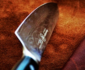 A close-up view of a large, sharp kitchen knife with a polished blade featuring intricate patterns. The handle is partially visible and appears to be made of a dark material. The background consists of a textured brown surface, possibly leather or fabric.