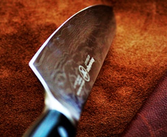 A close-up view of a large, sharp kitchen knife with a polished blade featuring intricate patterns. The handle is partially visible and appears to be made of a dark material. The background consists of a textured brown surface, possibly leather or fabric.
