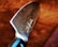 A close-up view of a large, sharp kitchen knife with a polished blade featuring intricate patterns. The handle is partially visible and appears to be made of a dark material. The background consists of a textured brown surface, possibly leather or fabric.