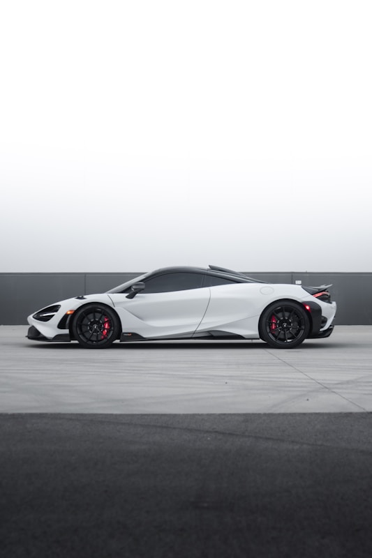 A sleek, modern white sports car with black wheels and red brake calipers is parked on a concrete surface against a minimalist gray and white background. The low profile and aerodynamic design emphasize speed and luxury.