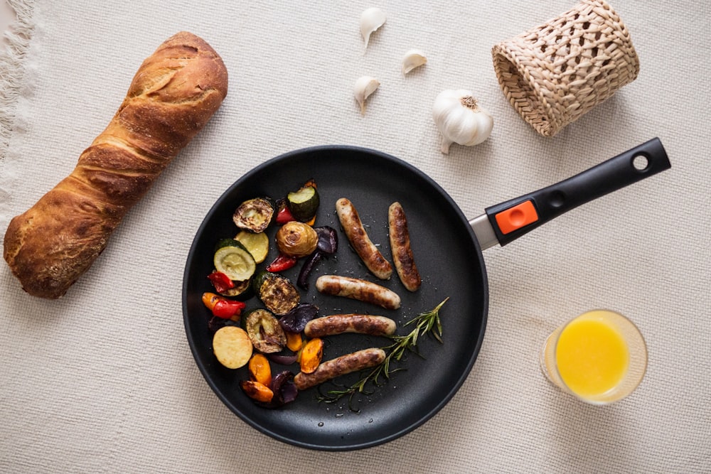 a pan filled with sausages and vegetables next to a glass of orange juice