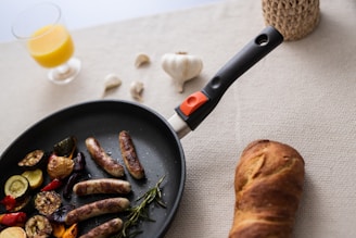 A frying pan containing grilled sausages and vegetables like zucchini, red peppers, and mushrooms is set on a textured surface. A loaf of crusty bread lies nearby. In the background, there are cloves of garlic scattered along with a woven basket and a glass of orange juice.