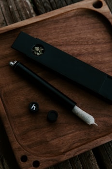 A neatly arranged set on a wooden tray featuring a black rectangular container, a smoking accessory resembling a joint, and a couple of small black lids. The container appears to hold some herbs.