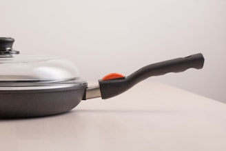 A frying pan with a glass lid rests on a light-colored surface. The pan has a black handle with a red element near its base, and the lid is slightly reflective.