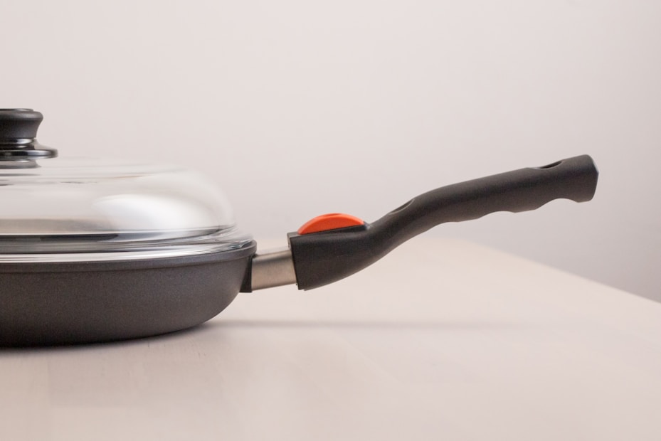 A frying pan with a glass lid rests on a light-colored surface. The pan has a black handle with a red element near its base, and the lid is slightly reflective.