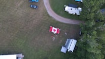 An aerial view captures a grassy area with a central campsite featuring a large Canadian flag laid out on the ground. Several campers and vehicles are parked nearby, and a fire pit is surrounded by people sitting. The scene is bordered by trees, and various items like chairs and tables are spread out in the area.