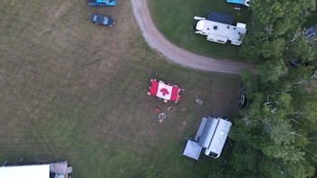 An aerial view captures a grassy area with a central campsite featuring a large Canadian flag laid out on the ground. Several campers and vehicles are parked nearby, and a fire pit is surrounded by people sitting. The scene is bordered by trees, and various items like chairs and tables are spread out in the area.