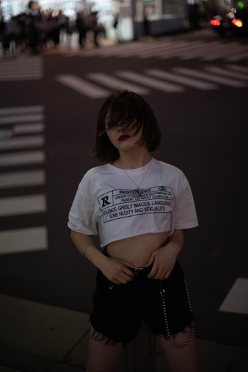 A person stands in an urban setting, likely at night. The individual wears a cropped white t-shirt with text on it, dark shorts, and a necklace. Their hair partially covers their face. The background features a street with a crosswalk and blurry figures in the distance, suggesting movement and possibly a busy intersection.