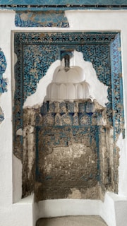 Ornate architectural detail featuring intricate blue and white tile work, interlaced with weathered patterns and exposed stone. The design reflects a historical aesthetic with geometric motifs and a partially deteriorated surface.