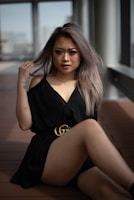 A woman with long, light brown hair sits on a wooden floor in a modern indoor space, wearing a black dress with a gold belt. She gazes directly at the camera with a confident expression, lightly holding her hair with one hand. Large windows in the background allow natural light to softly illuminate the scene.
